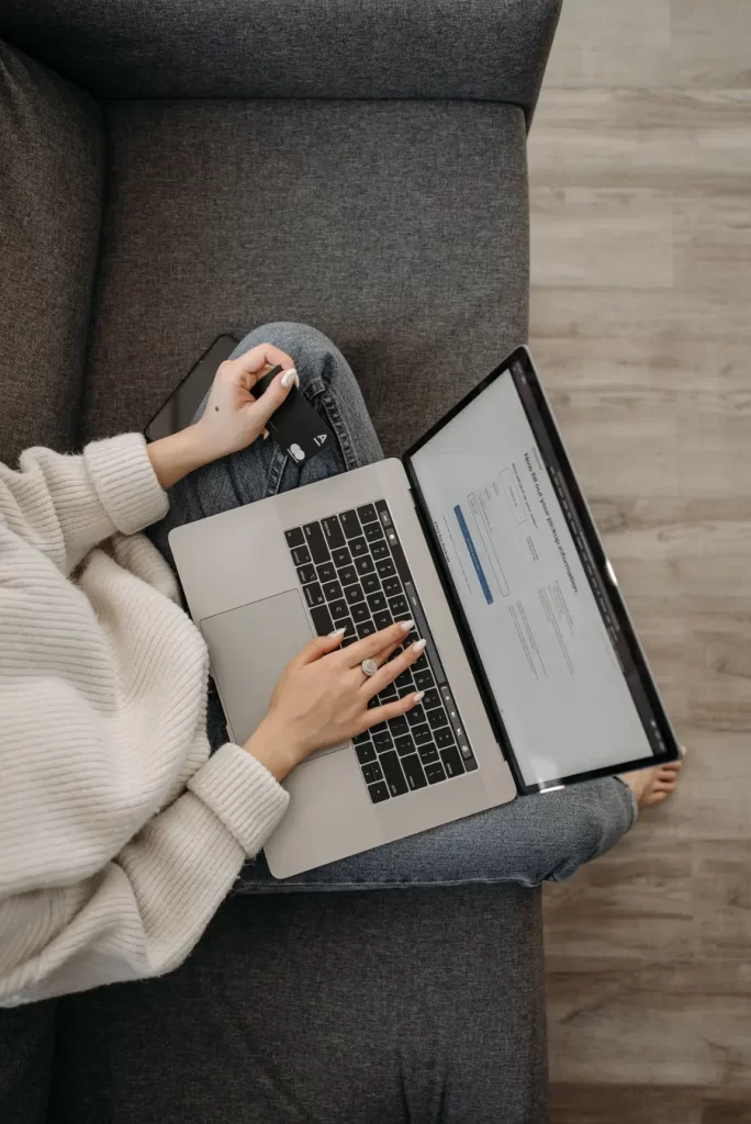 Top view of a woman shopping online using a laptop while sitting on a grey sofa. Image: Pexels/ Pavel Danilyuk