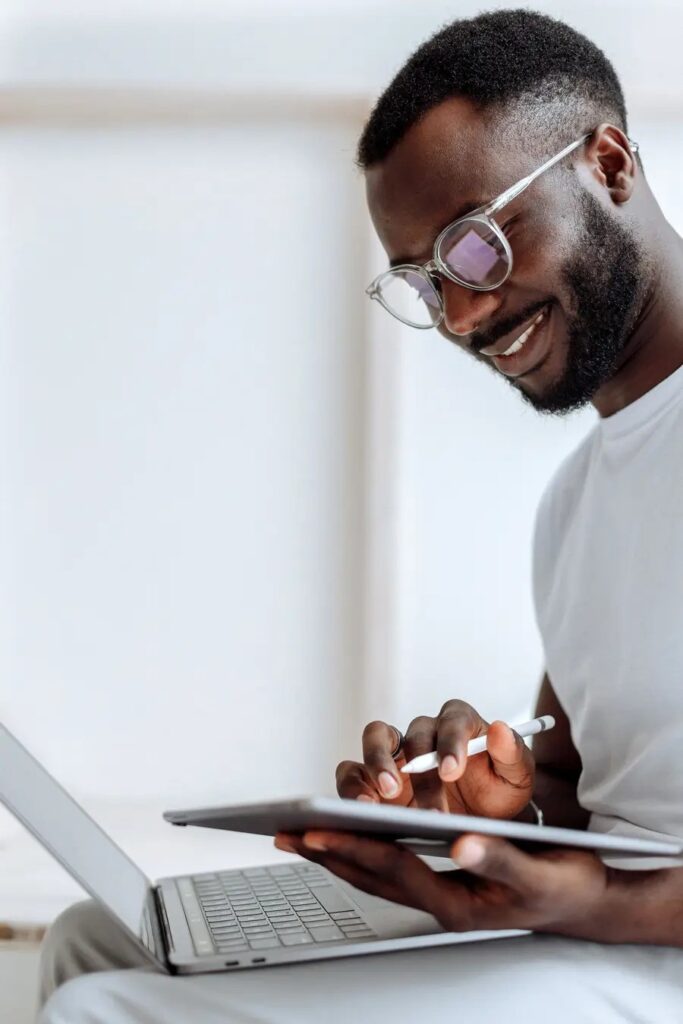 Image of an african man smiling. He is looking at a tablet and there i a laptop on the desk. Image: Pexels/ Thirdman